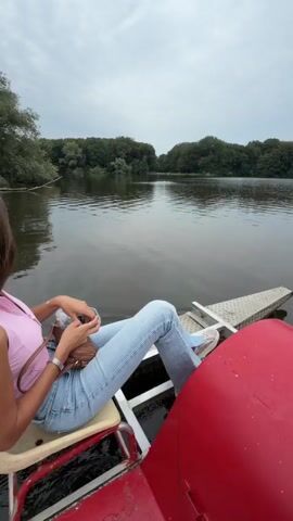 The video shows a first-person view of someone riding a pedal boat on a lake. The boat has red accents and metal platforms for the pedals. Trees and a cloudy sky are visible in the background. At 00:00, a person's legs in blue jeans and a brown jacket are visible. At 00:01, the person's feet in white sneakers and gray pants are shown on the pedals. At 00:02, the view shifts to show a person in a gray t-shirt looking up at the sky, with trees and the lake in the background. Several red buoys are visible on the water. At 00:03, the camera pans across the lake, showing a line of white buoys.