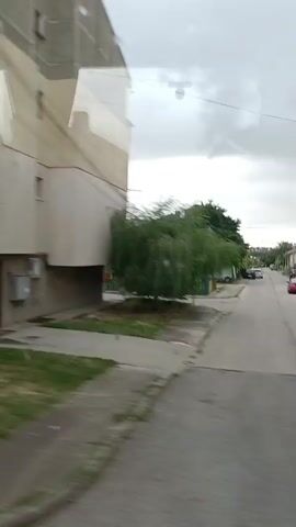 The video shows a blurry, panning view of an apartment building and its surroundings. The building appears to be made of concrete and has multiple stories with balconies and windows. There are some trees and grassy areas visible. The footage is from a moving vehicle, indicated by the motion blur. There are no people or distinct actions captured due to the low quality and speed of the recording.