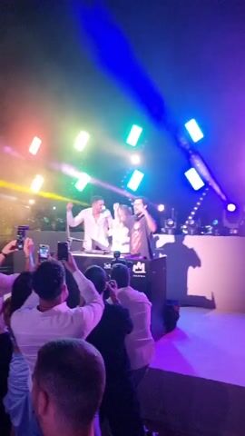 This video shows a nighttime outdoor event with a stage setup. Two individuals are visible behind DJ equipment, one in a white shirt and the other in a dark shirt. They appear to be performing or interacting with the audience. The stage is illuminated by colorful spotlights, including purple, blue, yellow, and green lights, with a smoky or hazy atmosphere. Several people are in the foreground, facing the stage and holding up smartphones, presumably to record or take pictures. The overall scene suggests a concert or party atmosphere with a DJ performance.