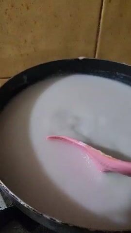 A close-up shot shows a pink slotted spoon stirring a white liquid in a black pan. The pan is on a stovetop, and the background is a tiled wall. The liquid appears to be simmering or cooking. The spoon is actively moving the liquid, creating ripples and small waves. The lighting is dim, suggesting an indoor setting. The focus is on the spoon and the liquid, with the background slightly blurred.