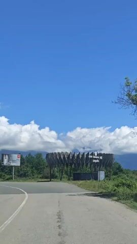 The video shows a daytime drive along a paved road in a rural or semi-rural setting. The sky is bright blue with large white cumulus clouds hovering low in the distance, partially obscuring mountains.

The camera moves forward, following a road that curves to the left. On the left side of the road, there is a billboard advertising what appears to be coffee, with the text "AMERI" and other partially visible letters. Green trees and dense foliage line both sides of the road and extend into the background.

On the right side of the road, a distinctive, large, dark-colored structure stands. It has a geometric, corrugated, or ribbed design, appearing to be a monument or an artistic gateway feature. The structure is made of what looks like metal or concrete, with several vertical supports. The overall scene suggests a journey through a scenic area, possibly entering or leaving a particular region. The location is specified as Inkiti, Abkhazia.