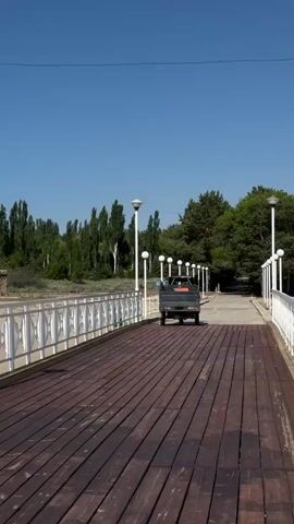 A dark colored pickup truck is slowly driving down a wooden boardwalk. The boardwalk has white railings on both sides and is lined with streetlights. Beyond the boardwalk, there is a body of water and a treeline with various types of trees, including tall, slender trees and wider pine trees. The sky is clear and blue, indicating a sunny day.