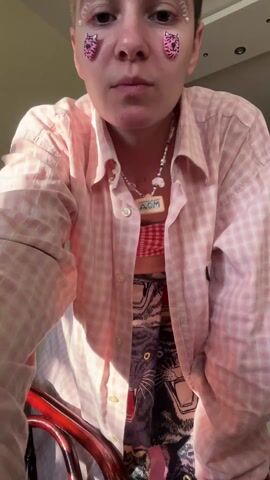 A person with short hair and heart stickers on their face dances in a room. The person is wearing a pink and white checkered shirt, a red and white checkered bra top, and a skirt printed with tiger faces. They move their arms and body to the music. The room is bright with sunlight. A red chair is in the foreground, and a cabinet is visible in the background. A doorway leads to another room.