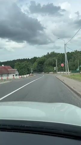 The video shows a view from the interior of a vehicle driving down a paved road. The road curves to the left and is bordered by a white and red striped barrier on the left and a wooden fence and grass on the right. Trees and vegetation line both sides of the road, with a few houses visible on the left. The sky is overcast with dark clouds. There are traffic signs visible, including a speed limit sign indicating 50. A circular speed limit marking of 50 is painted on the road.