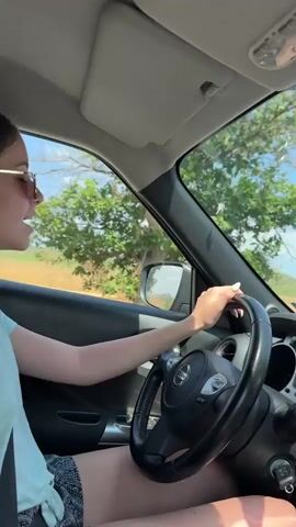 The video shows a young woman driving a car. She is wearing sunglasses, a light blue t-shirt, and patterned shorts. She has her right hand on the steering wheel and appears to be looking ahead. The interior of the car is visible, including the steering wheel with a Nissan logo, the dashboard, and part of the passenger seat. The exterior view through the windshield shows a sunny day with blue sky, white clouds, green fields, and trees. The camera appears to be positioned inside the car, to the right of the driver. The video is short, about 3 seconds long.