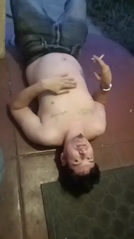 The video depicts a shirtless man wearing jeans lying on a tile floor. He has tattoos on his chest and arm, is holding a cigarette in one hand, and has his other hand resting on his stomach. A black mat is visible near his head. The lighting is subdued, with a mix of natural and artificial light sources. There is a door opening sound in the background.
