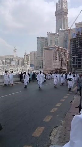 The video shows a street scene in Mecca, Saudi Arabia. Numerous people dressed in white ihram garments are walking on the side of the road. In the background is the Masjid al-Haram, the holiest mosque in Islam, as well as the Abraj Al-Bait Towers. A young boy stands by the side of the road looking at a mobile phone. The weather appears sunny with some clouds. Construction is visible on some buildings.