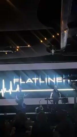 A dimly lit, indoor setting shows a band called Flatline performing on a stage. The stage has a white backdrop displaying the band's name in large, glowing white letters with a blue heart rate monitor graphic. The band consists of two men: one playing a bass guitar and the other singing and playing a keyboard. A saxophone stands nearby. String lights hang above the stage. In the foreground, the backs of several people face the stage, watching the performance. The camera briefly moves during the video, and fingers partially obstruct the view at times.