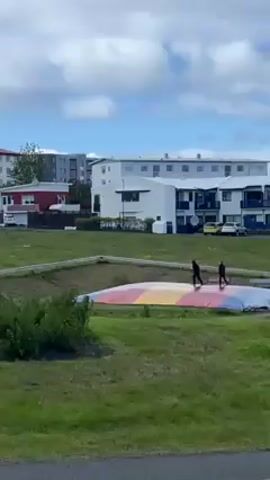 Here is a concise description of the video:

The video shows two people standing on a large, colorful, slightly oval-shaped trampoline set into a grassy field. The trampoline has various sections colored yellow, orange, and blue. The people are dressed in dark clothing and appear to be interacting with each other. Behind them is a neighborhood with houses, apartment buildings, and parked cars. The sky is partly cloudy, with patches of blue visible. The camera appears to be recording from a distance, across a road. As the video progresses, the trampoline's color scheme shifts due to lighting and possibly effects, appearing white and almost glowing. The surrounding greenery and urban landscape remain consistent.