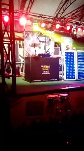 The video shows a stage setup for a musical performance. There’s a black DJ booth with the logo "VEEX BENZ DRUMZ ONEMANSQUAD" on it. Behind the booth is a set of drums. To the right of the booth is a large, white, rectangular structure with an array of LED lights that display changing patterns. A person wearing a white shirt stands behind the booth and appears to be operating the equipment. In the foreground, there are two conga drums. The stage is illuminated with red spotlights and various other colored lights. The stage is covered with a green rug and is situated beneath a metal frame structure, possibly a tent.