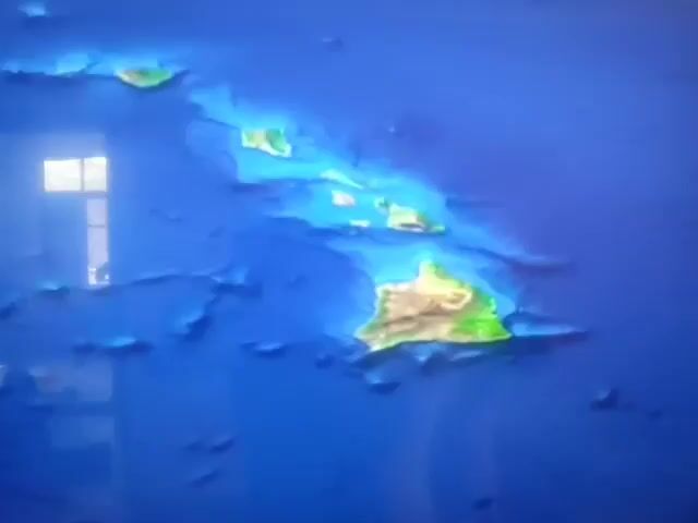 The video displays a 3D topographic map of the Hawaiian Islands. The islands are arranged in a chain, shown in shades of green and brown, with the surrounding ocean depicted in blue. A text label appears and hovers over the largest island, identifying it as "Hawaii." A small circular icon with colored dots briefly appears below the "Hawaii" label. The map appears to be a digital display, with a reflection of a window visible on the left side of the screen.