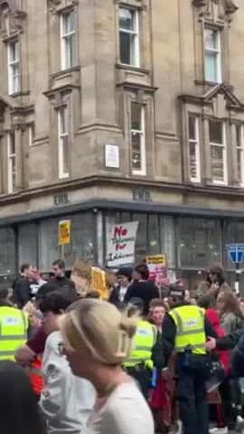 The video shows a crowd of people gathered on a city street, in front of a building with "END." written on its corner. Some people are holding signs, one of which reads "No Tolerance For Intolerance." Several individuals wearing yellow reflective vests appear to be providing security or maintaining order. The environment suggests a demonstration or public gathering, with the crowd likely protesting or advocating for a specific cause. The people in the crowd are diverse, with different ages, genders, and clothing styles. The architecture of the building is urban, with a classic stone facade and large windows.