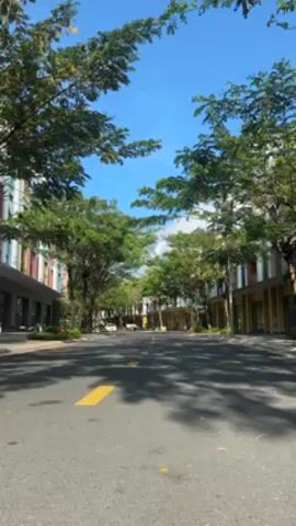 The video shows a wide, empty street lined with trees and buildings with colorful facades. The street has yellow markings and a few parked cars are visible in the distance. The sky is blue with some clouds, and the trees provide a green canopy over the street. A child's voice is heard in the background.