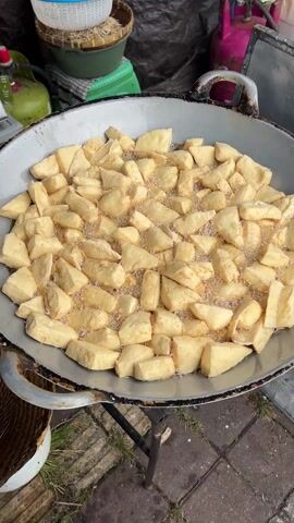 A large metal wok is filled with small, irregularly shaped pieces of light tan tofu, immersed in bubbling oil. The tofu is actively being fried, as evidenced by the bubbles forming around each piece. The wok is supported by a simple metal stand, placed on a paved surface with patches of green grass visible in the cracks. Behind the wok, out of focus, are several items, including a basket, a green canister, and a pink propane tank. The focus is sharply on the tofu and the frying process within the wok.