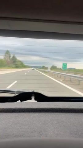 The video, captured from inside a car, presents a view through the rear window of a highway. The sky is overcast, and the road appears to have multiple lanes with sparse traffic. A large truck travels in the right lane. Greenery lines the side of the highway. The rear window has horizontal heating wires and a windshield wiper arm. A dark grey, fabric-covered surface occupies the lower portion of the frame, indicating the car's interior.
