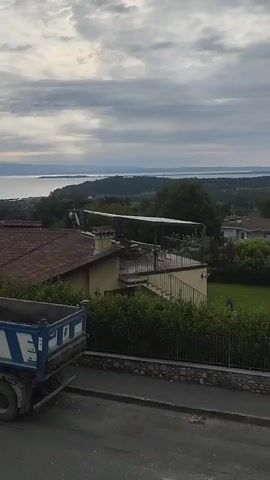A dump truck with a blue and white striped bed is parked on a paved road in front of a residential area. Beyond the houses, a large body of water is visible, with hills or mountains in the distance. The sky is cloudy.
