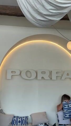 This media file features an interior scene with a woman sitting on a couch in the foreground. She is partially visible, with her back to the camera, wearing a white tank top and jeans. Her blonde hair is down. She appears to be engaged with something off-camera.

The background is dominated by a large, circular white wall with the word "PORFA" spelled out in large, white, three-dimensional letters. The letters are evenly spaced and cast subtle shadows on the wall. An illuminated, golden-toned strip light curves around the upper portion of the circle, creating a halo effect. Above this, exposed wooden beams are visible, suggesting an older architectural element. Draped fabric, possibly a curtain or decorative element, hangs from the ceiling.

The overall aesthetic is modern and minimalist, with a warm and inviting ambiance, likely due to the lighting. The pillows on the couch in the foreground add a touch of comfort and texture, with one featuring a blue and white geometric pattern.

The scene appears to be set indoors during the daytime, with bright lighting. The setting is likely a cafe, restaurant, or a boutique establishment given the prominent branding and stylish decor. The woman's posture and relaxed demeanor suggest a casual and comfortable atmosphere. The visible text "PORFA" is the most prominent detail.
