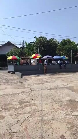 The video shows an outdoor area on a sunny day. In the foreground, there is a paved area with some discolored patches. In the middle ground, there is a raised concrete seating area with several people sitting and standing, some under colorful umbrellas. Behind them, there are trees and buildings. To the right, there is a tall, modern building with multiple stories and windows. Further to the left, there is a single-story building with a tiled roof and an entrance that says "MADRASAH IBTIDAIYAH MUHAMMADIYAH KARANGLEWAS". Several motorcycles are parked in front of the buildings. In the background, there are more trees and buildings, and the sky is clear blue.