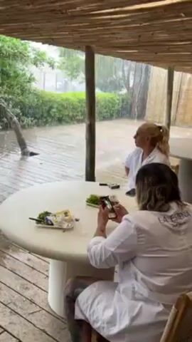 Two individuals are under a partially covered area with a bamboo ceiling, likely a patio or outdoor seating space, during a heavy rainstorm. Both are wearing white lab coats. One individual, with long hair and tattoos on their arm, is holding a smartphone in a vertical orientation and appears to be taking a photo or video of the other person, who is looking at the camera and smiling. There is a white, round table between them with what seems to be unfinished food and items like napkins and containers. The backdrop is a lush green area, visible through the downpour of rain. The ground outside is flooded with water. The words "LA ESTERNA" are embroidered on the back of the person taking the picture's lab coat.