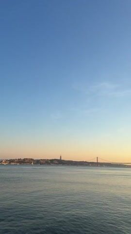 A wide shot captures a body of water under a clear, pale blue sky that transitions to a soft orange hue near the horizon. In the distance, a city skyline is visible, punctuated by a tall, slender monument. A suspension bridge spans across the water, connecting distant landmasses. The water's surface is rippled, reflecting the subtle colors of the sky. The video progresses slightly to reveal a woman with dark hair, wearing a purple cap and a pink top, seated near the edge of the water, looking down. She is positioned in the lower right quadrant of the frame. The overall atmosphere is calm and serene, suggesting either dawn or dusk.