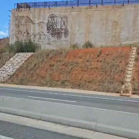 A vehicle is driving past a concrete retaining wall on the right side of the road. The retaining wall has a mural depicting horses and a wagon. The wall is covered in a light-colored concrete and the surrounding ground is covered in reddish-brown dirt and vegetation. There is a metal fence at the top of the retaining wall. There is a concrete barrier in front of the vehicle. The sky is blue with some clouds.