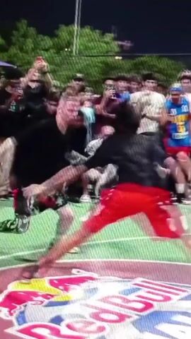 The video shows a freestyle soccer performance taking place at an outdoor event at night, surrounded by a crowd. The performance occurs on a marked area with the "Red Bull" logo prominently displayed. One performer, dressed in a black shirt and shorts with colorful patterns, executes tricks with a soccer ball. The crowd cheers and records the performance with their phones. The scene is illuminated by overhead streetlights.