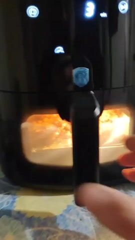 A person pulls the drawer of a black air fryer open to reveal two cooked patties sitting on parchment paper. The air fryer has a digital display showing numbers. The surface beneath the fryer is a blue and white floral pattern.