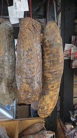 The video shows a display of cured meats hanging from a rack. The meats are of various sizes and shapes, and many have a moldy exterior. There are also some sliced meats in plastic packaging on a table below. A meat slicer is visible on the table. There are price tags attached to some of the hanging meats. In the background, a person in a green shirt is partially visible, and another person wearing a light-colored jacket appears to be behind the counter. The environment seems to be a market or a food shop.