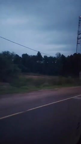 The video shows a dark, cloudy sky, likely dusk or dawn. A tall telecommunications tower with red lights is visible on the right side of the frame, behind a line of dark trees. A road with a yellow dividing line runs across the bottom of the frame. A white vehicle is blurred as it passes from left to right, its taillights and headlights visible. Another vehicle, possibly a silver SUV, is also blurred as it passes in the opposite direction. The scene suggests a rural or semi-rural roadside environment.