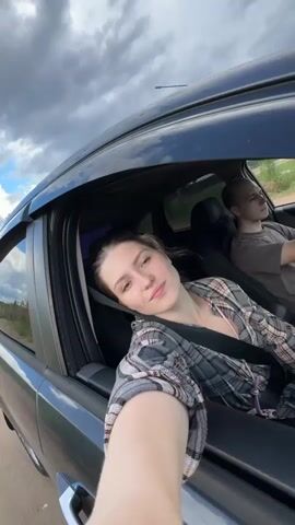A woman with her eyes closed is leaning out of a car window with her head resting on her arm, her hand holding the camera. She is wearing a plaid shirt over a dark top and a seatbelt. In the background, a man is visible in the driver's seat, looking ahead. The car is moving, and the environment outside appears to be rural with trees and a cloudy sky.