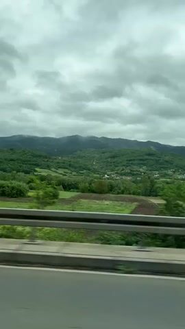 The video shows a view from a car moving along a road. The weather is cloudy. There is a safety rail and some greenery alongside the road. Beyond that are trees and agricultural fields, with forested hills visible in the distance. There is no sign of people in the view.