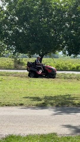 A man is seen operating a red and black riding lawn mower on a sunny day in Kran, Bulgaria. He is mowing a grassy area that runs parallel to a paved road, which is visible in the foreground. The man is wearing a baseball cap, a dark short-sleeved shirt with white stripes on the sleeves, and dark pants. He is seated on the mower, driving it from left to right across the frame, cutting the grass. Behind the grassy strip and the mower, there are several large trees casting shadows, and beyond them, a wide field or distant hills can be seen under a bright sky. The setting appears to be outdoors, likely in a rural or semi-rural area. No discernible text is visible on the man's clothing or the mower.