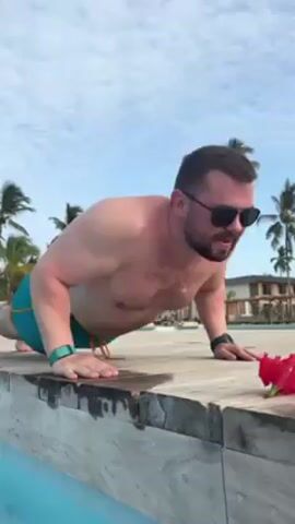 The video shows a shirtless man with a beard and sunglasses doing push-ups on a wooden ledge by a pool. He is wearing turquoise swimming trunks and two smart watches. A red flower sits on the ledge next to him. Palm trees and a building are visible in the background under a partly cloudy sky.