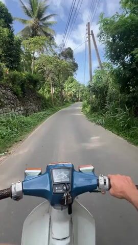 A first-person view shows a person riding a light blue and white vintage Honda scooter on a narrow paved road lined with lush tropical vegetation. The sky is blue with scattered clouds, and utility lines run overhead.  The scooter's dashboard is visible, showing the speedometer and fuel gauge. The rider's hand is on the right handlebar, controlling the throttle. Small houses and structures are intermittently visible through the trees along the roadside.