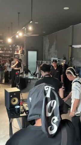 The video shows a group of people in what appears to be a retail store or studio space. In the foreground, a person wearing a black hooded garment with white lettering is partially visible from behind, facing away from the camera. To the left, a black speaker sits on a black table next to a DJ setup with turntables and a mixer. A person in a black t-shirt and blue jeans stands behind the DJ equipment, wearing headphones. Another person in a black t-shirt stands to the right of the DJ, looking towards the DJ setup. Further in the background, several individuals are standing and observing. To the right of the DJ area, a person with blonde-braided hair is visible, and a large, metallic, spiky headpiece is positioned on a mannequin or stand behind them. The space has concrete walls, track lighting, and clothing racks with garments displayed. An air conditioning unit is mounted high on the wall. The overall impression is of a creative or promotional event taking place.