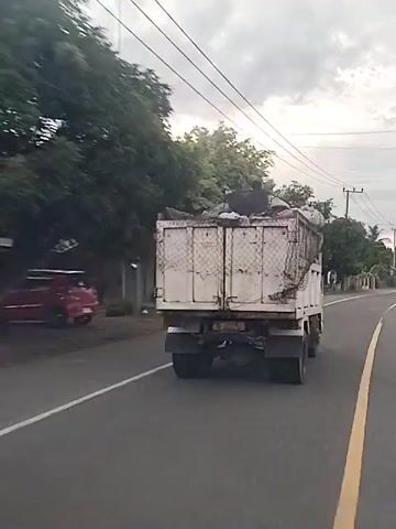 A video shows a white dump truck, heavily loaded, driving down a road. The truck is old and rusty, with the bed filled with what appears to be trash or discarded items. A person is sitting atop the load. The truck has a mesh net partially covering the back to secure the load. It moves from left to right across the screen, revealing a rural or semi-urban setting with trees, power lines, and a few buildings in the background. The sky is overcast.