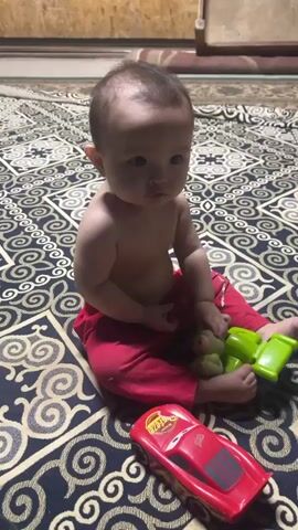 A baby is sitting on a patterned rug, playing with toys. The baby, wearing red pants and no shirt, has dark hair and is looking at the camera. To the baby's right is a green toy, and in front of the baby is a red toy car resembling Lightning McQueen. The scene appears to be indoors, with a wooden background visible at the top of the frame. The lighting suggests it could be daytime. The baby's expression is neutral. The pattern on the rug is intricate, featuring swirling black and gold designs.
