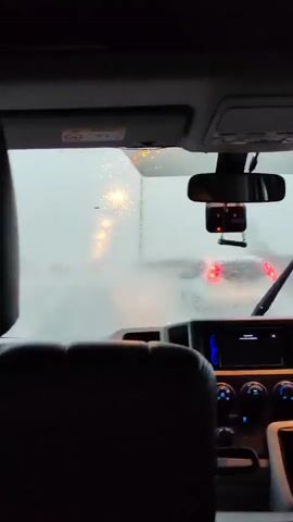 The video shows a view from inside a vehicle, looking through the windshield. It is raining, and the windshield wipers are active, clearing the water. Outside, other vehicles are visible, their lights blurred by the rain and mist. The scene appears to be a road or a parking lot, with streetlights visible in the distance, casting a hazy glow through the inclement weather. The interior of the vehicle includes a dashboard with a radio and various controls. The overall atmosphere is gloomy due to the weather.