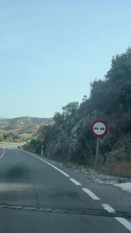 A video taken from a moving vehicle shows a winding road with a rocky mountainside on the right and a gentle slope of hills on the left. The sky is clear and blue. There is a guardrail along the side of the road and a few trees and bushes are visible. The road surface appears to be asphalt.