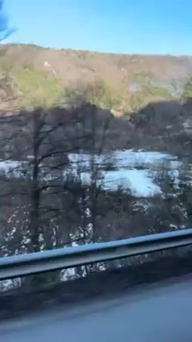 A vehicle is traveling on a road next to a body of water, possibly a river or lake, with ice or snow visible on its surface. Trees line the bank of the water, and a hill or small mountain is visible in the background. The sky is blue. The view is from inside the vehicle, looking out the side window. The road has a gray guardrail. The camera appears to be handheld. There is music playing.