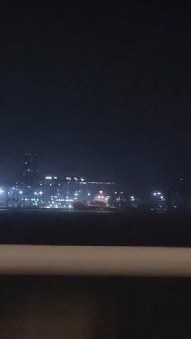The video shows a nighttime view of a brightly lit industrial area across a body of water. The lights appear to be from a port or industrial complex, with numerous tall structures, possibly cranes, visible. The sky is dark, and the water reflects some of the lights. A blurry foreground element, possibly a railing or barrier, partially obstructs the view. The overall impression is of a busy, active industrial site operating at night.