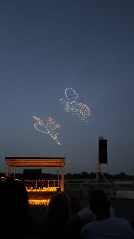 Here's a detailed description of the video:

The video shows an outdoor event at night, with a lit-up stage and a drone show in the sky. The sky is dark with a hint of twilight. Two formations of drones are illuminated, one resembling a horse and the other a jockey. 

Below the drone formations, there is a stage with people on it, backlit by many lit candles. The people on stage appear to be musicians or performers. In the foreground, the backs of the heads of some audience members are visible. To the right of the stage, there is a tall speaker tower. Beyond the audience and the stage, the outlines of trees and buildings can be discerned. The location appears to be a park or open area.