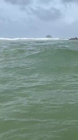The video shows a choppy, green ocean under a cloudy sky. Waves are breaking in the distance, and a small, rocky island with some trees on top is visible on the horizon. The water appears to be in motion, with ripples and whitecaps. There are no people or man-made objects in the immediate foreground.
