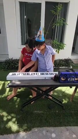 Two children are in an outdoor setting, possibly a yard, in front of a building with a large window. They are positioned around a black table on which a white keyboard and a blue portable speaker sit. The child on the left, wearing a red shirt, is seated and playing the keyboard. The child on the right, wearing a light blue button-down shirt and a blue party hat, is standing and observing. The child on the right appears to be looking at the keyboard and possibly singing or speaking. The ground is covered in artificial turf.