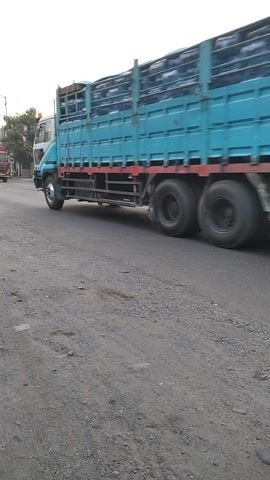 The video captures a busy street scene in Jubung, Indonesia, during what appears to be late afternoon or early evening, indicated by the soft, diffused sunlight and the warm glow on the horizon. The weather seems to be clear with some scattered clouds.

The primary subjects are various vehicles navigating the road. A large, light blue truck with a covered cargo bed is prominently featured, moving along the asphalt road. Alongside it, several motorcycles are also in motion. Some of these motorcycles carry multiple passengers, with riders and pillions often wearing helmets. One motorcycle is a bright red scooter, while others are white and black.

The setting is a roadside environment with buildings lining one side of the street. Some of these buildings appear to be shops or small businesses. One building has a sign that reads "RADIATOR RIzki". Flags are also visible, attached to poles or vehicles. The road surface is a mix of asphalt and gravel, suggesting it might be a main road in a more rural or developing area.

The overall activity is the movement of traffic. The vehicles are either moving or stopped, indicating a normal flow of transportation. The presence of multiple vehicles, including heavy trucks and numerous motorcycles, suggests a fairly active commercial or commuter route. There are no overt signs of distress or unusual events, suggesting a typical day on this Indonesian road. The atmosphere appears to be one of everyday activity and commuting.