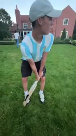 A young man wearing an Argentina jersey, shorts, a baseball cap, and sneakers is standing in a grassy yard with a cricket bat. He is holding the bat and appears to be preparing to swing. In the background, there is a large brick house and other people are visible in the distance. The man takes a practice swing with the bat. The sky is overcast.