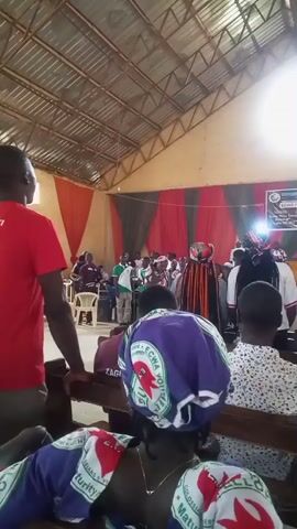 A video shows a gathering inside a building with a corrugated metal roof. In the foreground, several people are seated on wooden benches, facing away from the camera. They are wearing colorful headwraps. In the mid-ground, a group of people are standing, and some appear to be involved in a performance or ceremony. One individual is wearing a distinctive, elaborate costume with fringe. A banner with text and graphics is visible on the back wall. The overall lighting suggests an indoor event during the daytime.