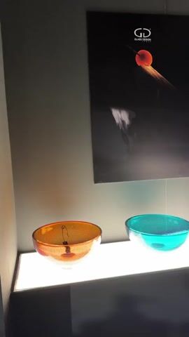 The video showcases an indoor display of various colorful glass bowls. The bowls, in shades of orange, turquoise, red, blue, grey, and clear, are arranged on a brightly lit white shelf, creating interesting light and shadow effects. Above the bowls, a dark poster features the "GD GLASS DESIGN" logo and an abstract image of an orange sphere with a trail. As the camera pans, Italian text "Disegnare la luce con forme e colori Designing l[ight]" is visible on the dark wall behind the display. The scene suggests an exhibition or retail environment, potentially related to glass art or design, in Rho, Italy.