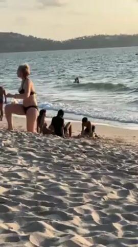 The video shows a beach scene with several people. A woman in a black bikini walks towards the left of the frame. Three people are sitting on the beach: a woman in a brown bikini, a person in a black shirt, and a child who is digging in the sand. Several people are playing in the water in the background. The sky is a pale yellow, suggesting it is either sunrise or sunset. There is a heavily wooded hill in the background. The sand in the foreground is uneven, with footprints and lines visible. The water is slightly choppy with small waves breaking on the shore.