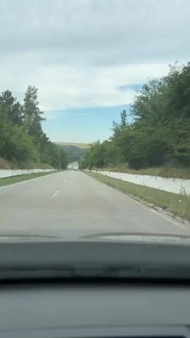 The video shows a first-person view from inside a vehicle traveling on a paved road. The road is bordered by trees on both sides. There are other vehicles on the road, including cars and what appears to be a truck in the distance. The sky is cloudy with patches of blue. The road surface is cracked in places, and there are white concrete barriers along the sides of the road. The landscape appears to be hilly, with fields visible in the distance. The video is shot in daylight.